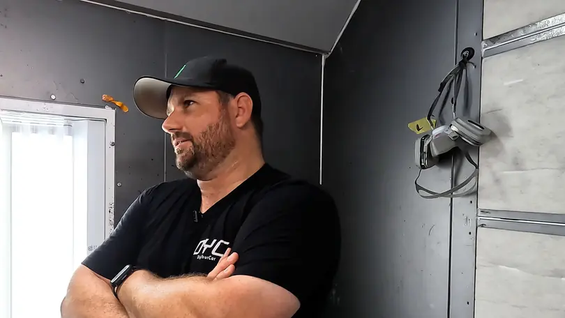 A presenter in a baseball cap, arms folded in the spray booth, smiles at someone off camera
