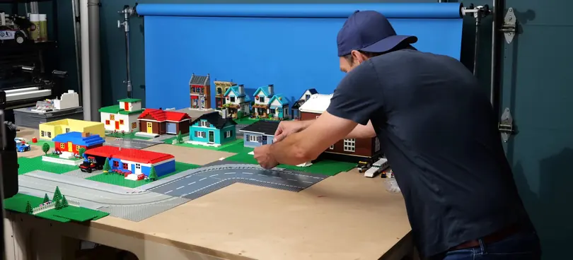 Andy Bailey lays a road piece into a LEGO town on a plywood bench in front of a blue paper backdrop