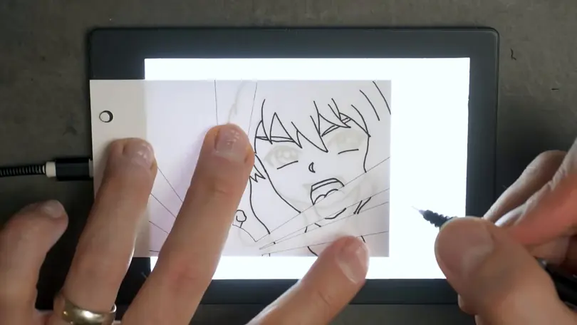 Fingers hold a flipbook page down on a light box while a pen inks the next drawing of a shouting face