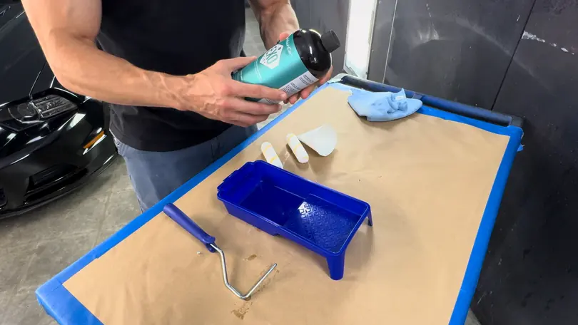Hands shake a bottle over a paper-lined bench with a paint tray and a roller, a black car beside it
