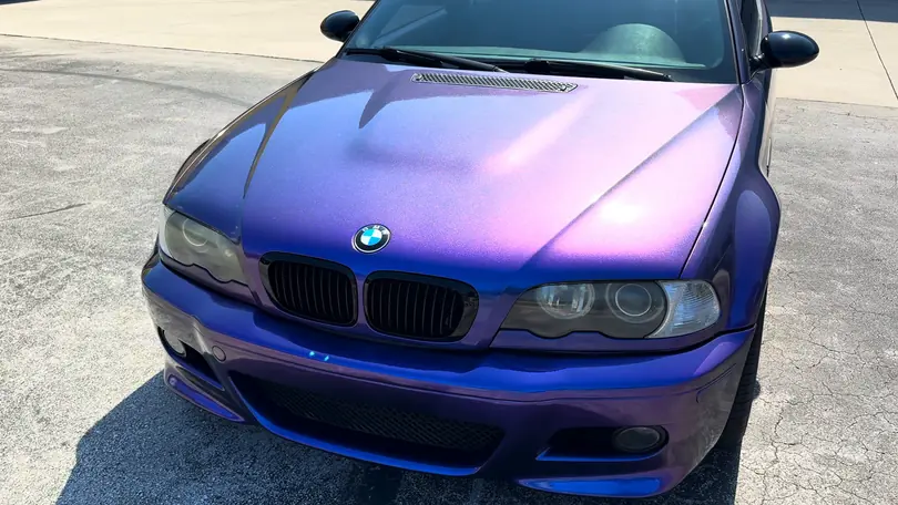 The front of a BMW M3 in a finish that shifts from violet to blue, parked in the sun