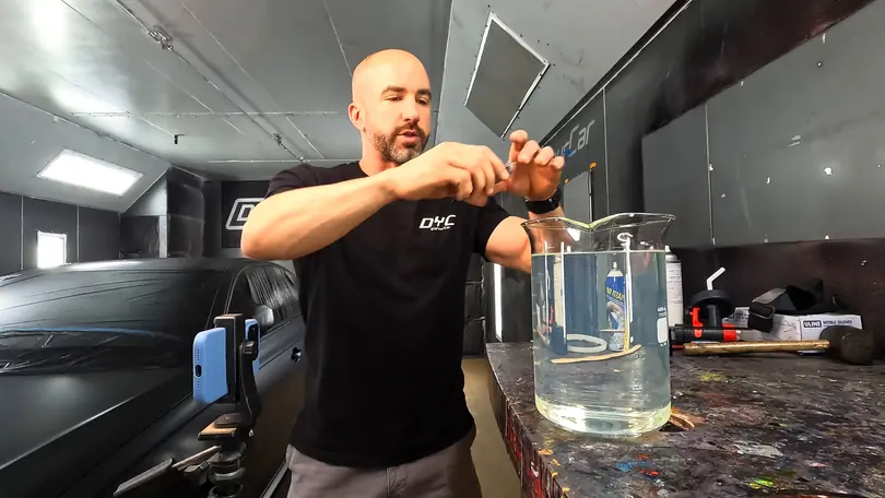 A presenter measures out a sample over a beaker at the bench of a spray booth, a phone on a stand filming beside him