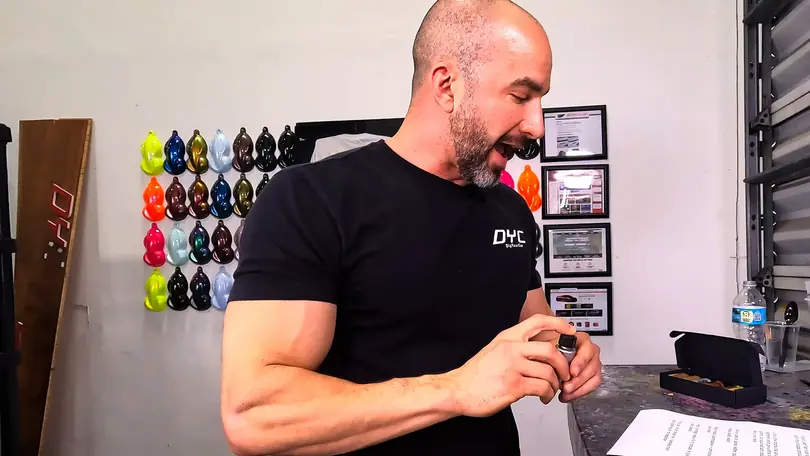 A presenter checks a small bottle at a workbench, a wall of painted color swatches behind him